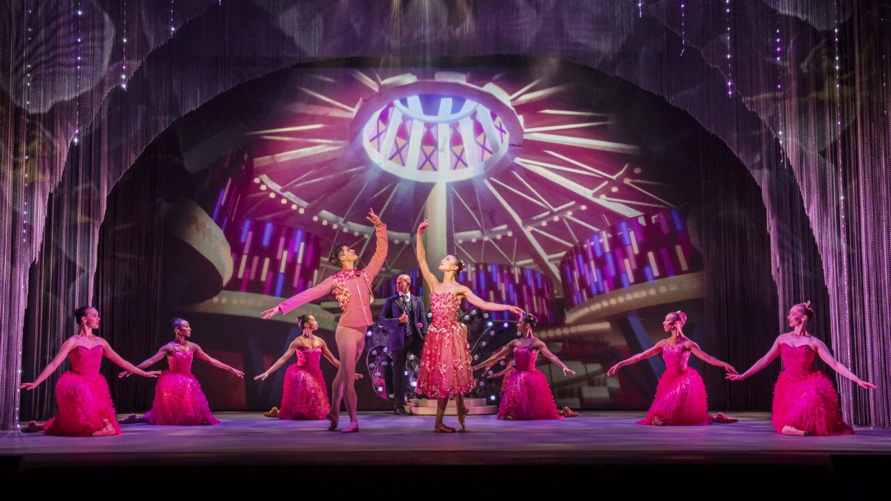 Dancers in bright pink costumes perform on stage with a dramatic circular light backdrop, while a man stands in the center under spotlights.