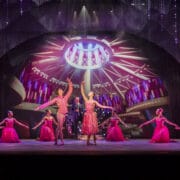 Dancers in bright pink costumes perform on stage with a dramatic circular light backdrop, while a man stands in the center under spotlights.
