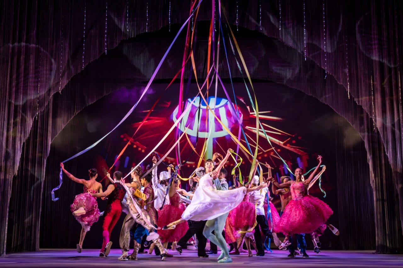 A group of performers dance energetically on stage, holding colorful ribbons with vibrant lighting and a dynamic background.