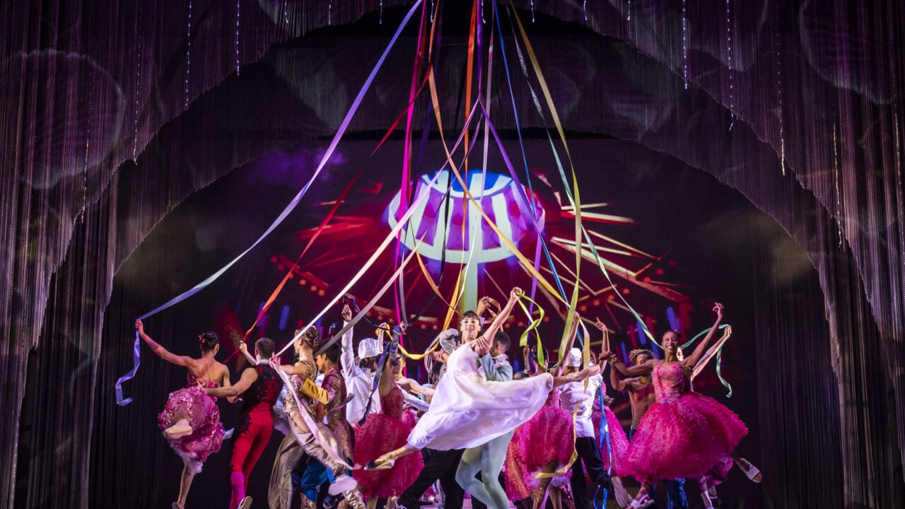 Performers in colorful costumes dance energetically on stage, holding long ribbons overhead, with vibrant lighting and a dynamic backdrop enhancing the festive atmosphere.
