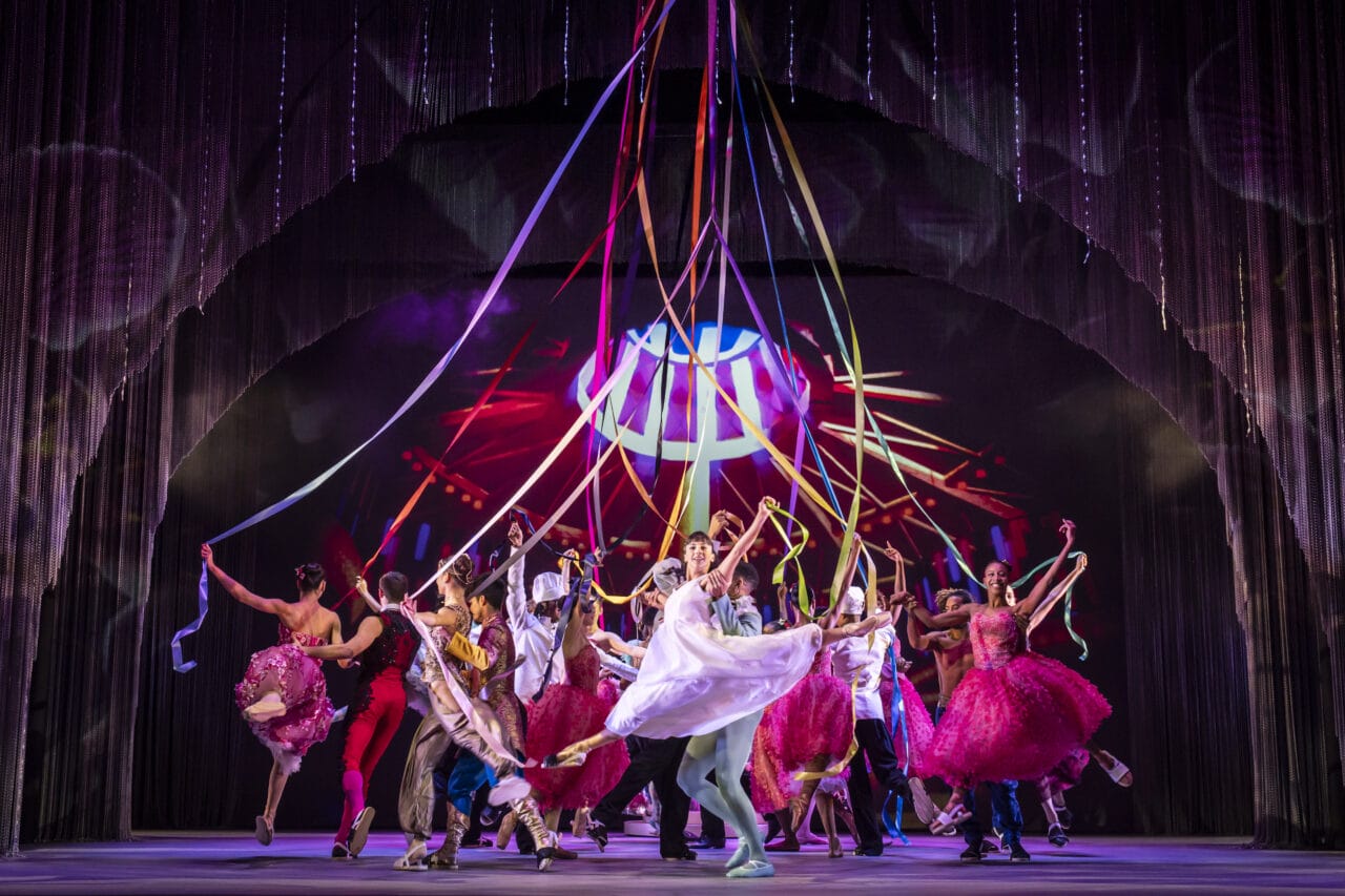 Performers in colorful costumes dance energetically on stage, holding long ribbons overhead, with vibrant lighting and a dynamic backdrop enhancing the festive atmosphere.