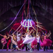 Performers in colorful costumes dance energetically on stage, holding long ribbons overhead, with vibrant lighting and a dynamic backdrop enhancing the festive atmosphere.