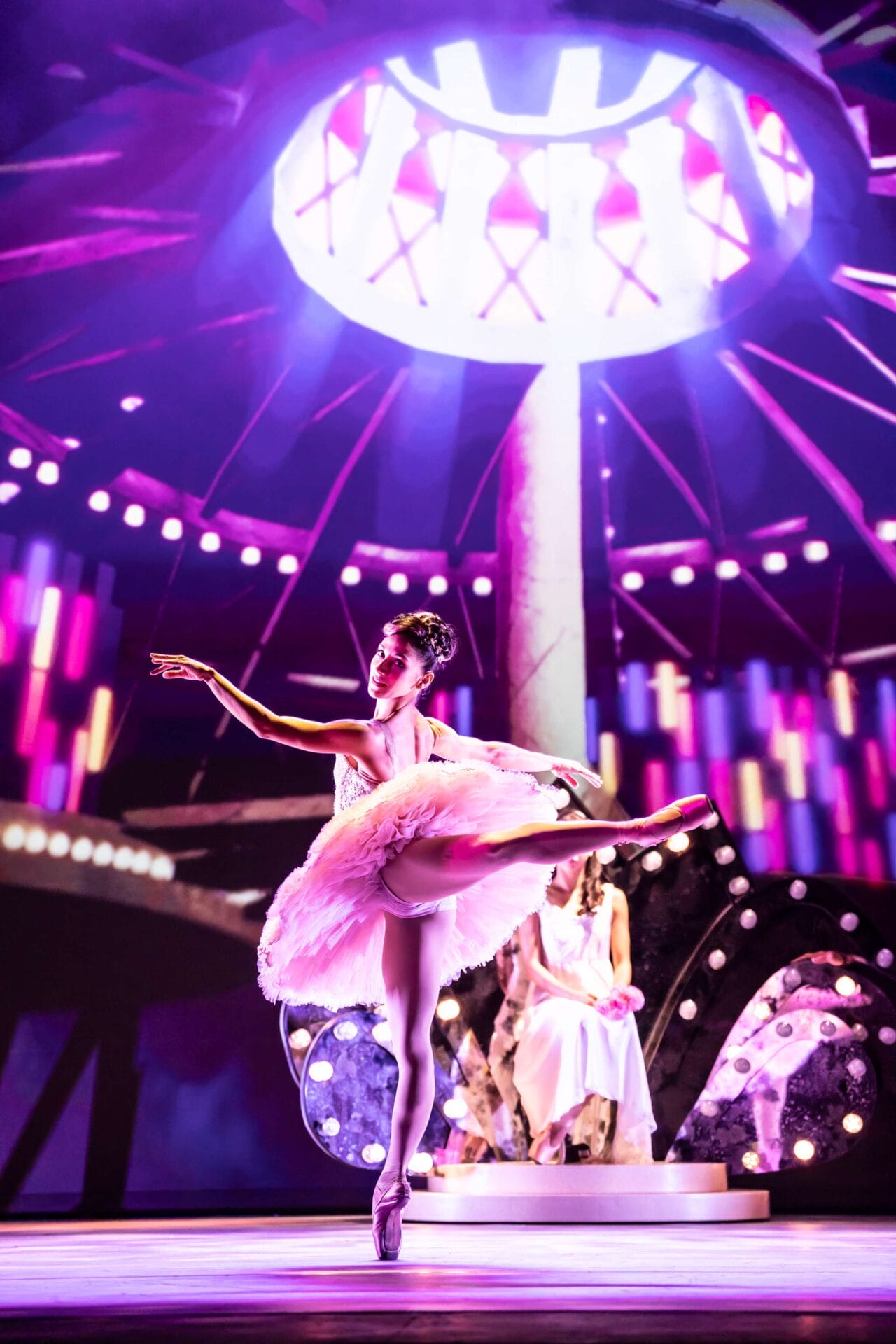 A ballerina in a pink tutu performs an arabesque on stage, with colorful lights and a seated figure in the background.