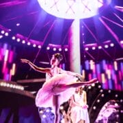 A ballerina in a pink tutu performs an arabesque on stage, with colorful lights and a seated figure in the background.