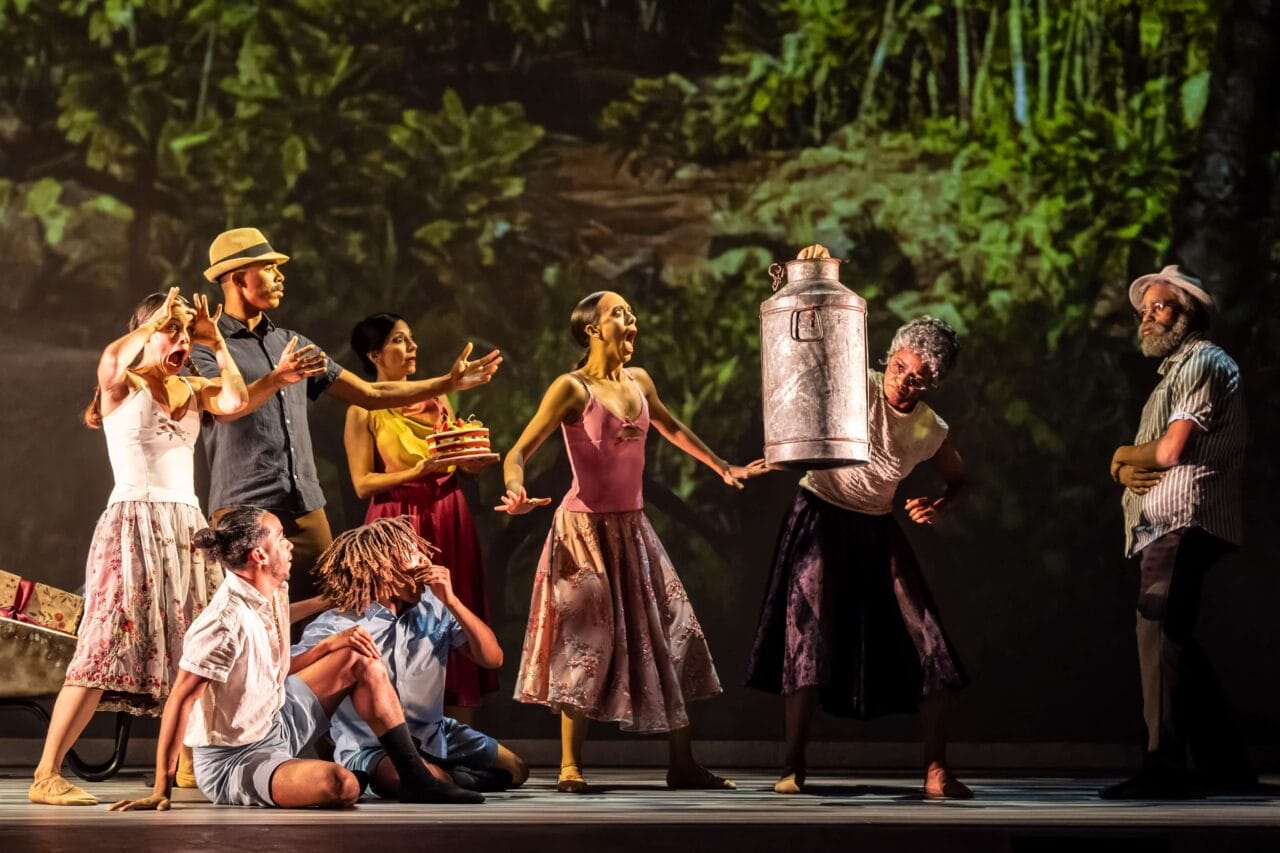 A group of performers on stage act out a lively scene; one person lifts a large metal canister while others react expressively, with a forest backdrop behind them.
