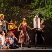 A group of performers on stage act out a lively scene; one person lifts a large metal canister while others react expressively, with a forest backdrop behind them.