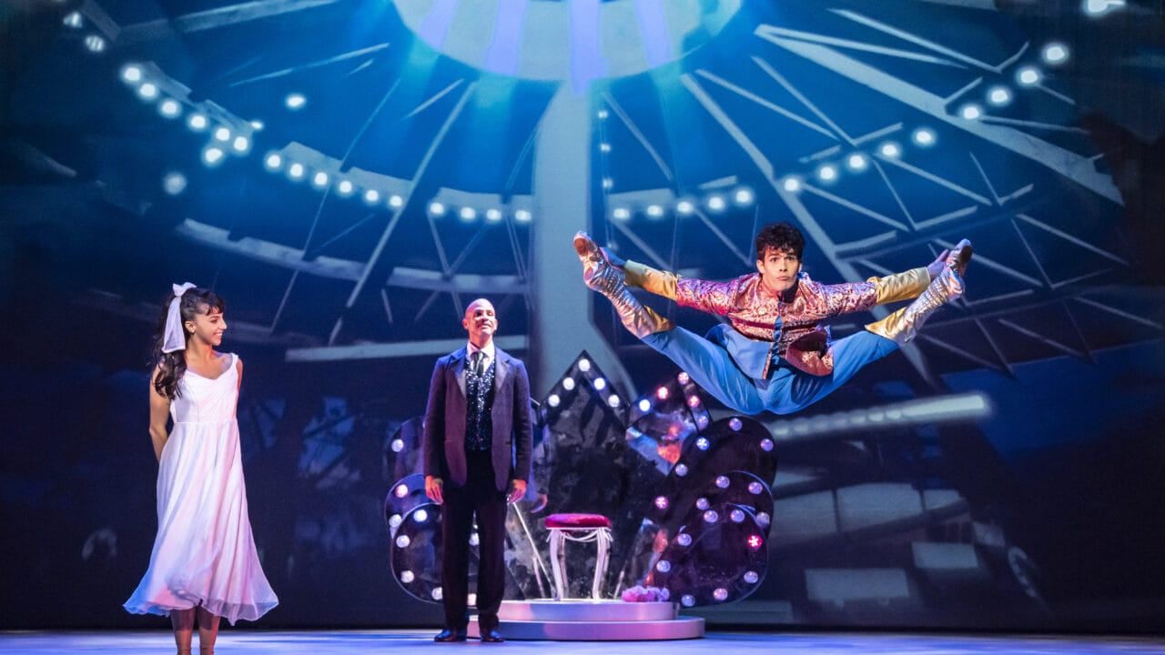 A performer in colorful attire jumps midair with legs extended, while a woman in a white dress and a man in a suit watch on a brightly lit stage.