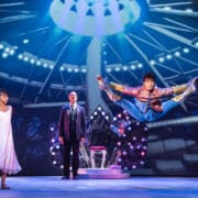 A performer in colorful attire jumps midair with legs extended, while a woman in a white dress and a man in a suit watch on a brightly lit stage.