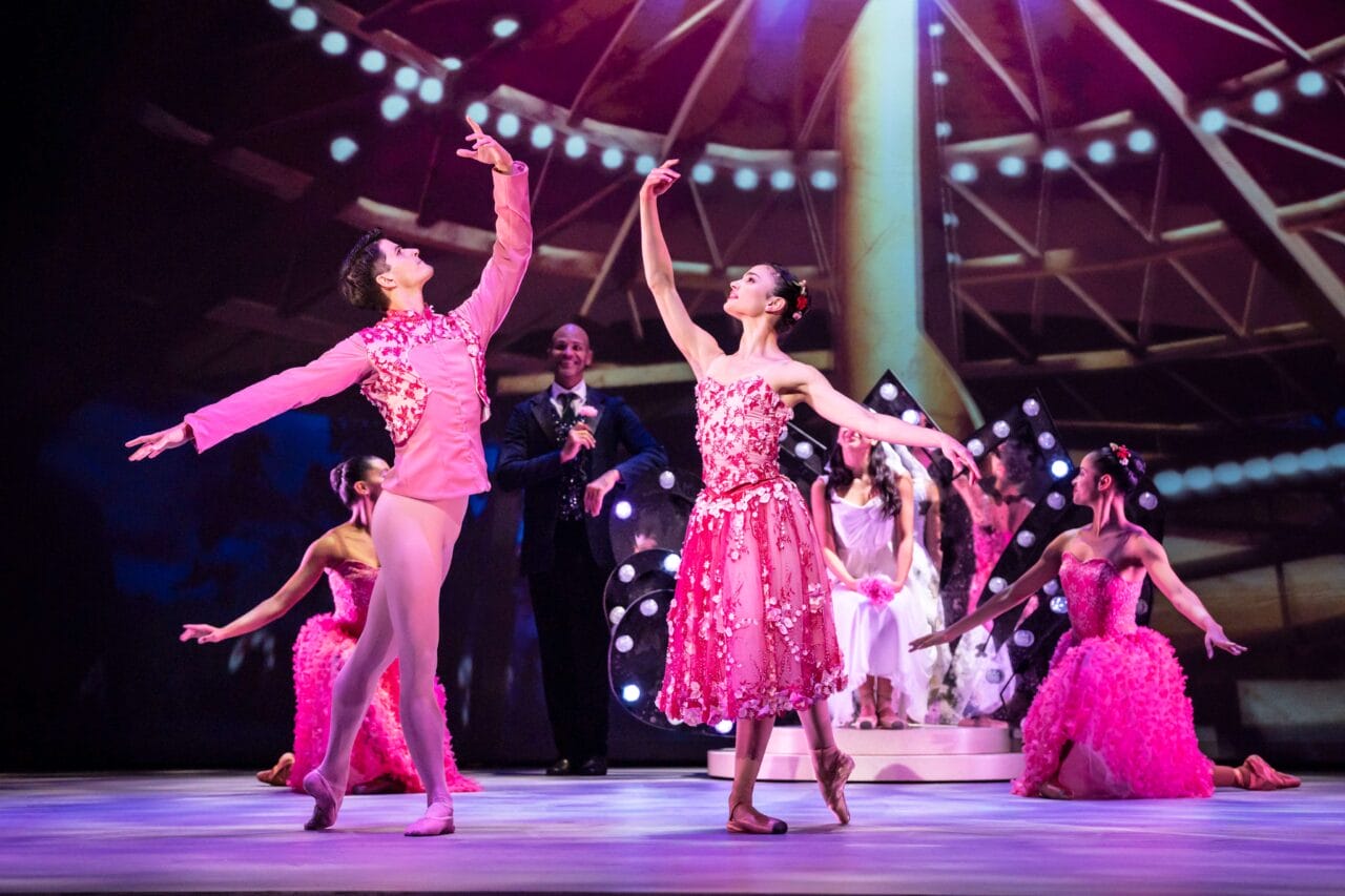 Ballet dancers in pink costumes perform on stage with bright lights and seated performers in the background.