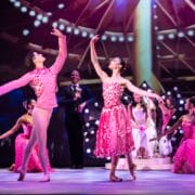 Ballet dancers in pink costumes perform on stage with bright lights and seated performers in the background.