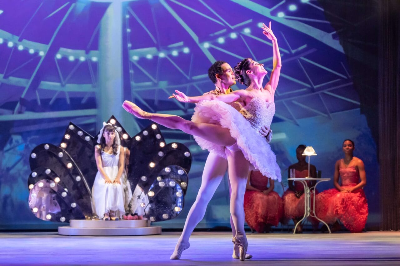 Two ballet dancers perform a lift on stage, while a seated woman and two others in pink costumes watch in the background under theatrical lighting.