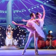 Two ballet dancers perform a lift on stage, while a seated woman and two others in pink costumes watch in the background under theatrical lighting.