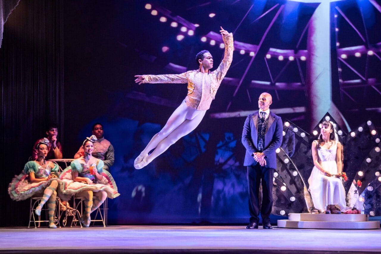 A ballet dancer leaps mid-air on stage while others, including people in costumes and formal wear, watch in the background.