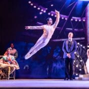 A ballet dancer leaps mid-air on stage while others, including people in costumes and formal wear, watch in the background.