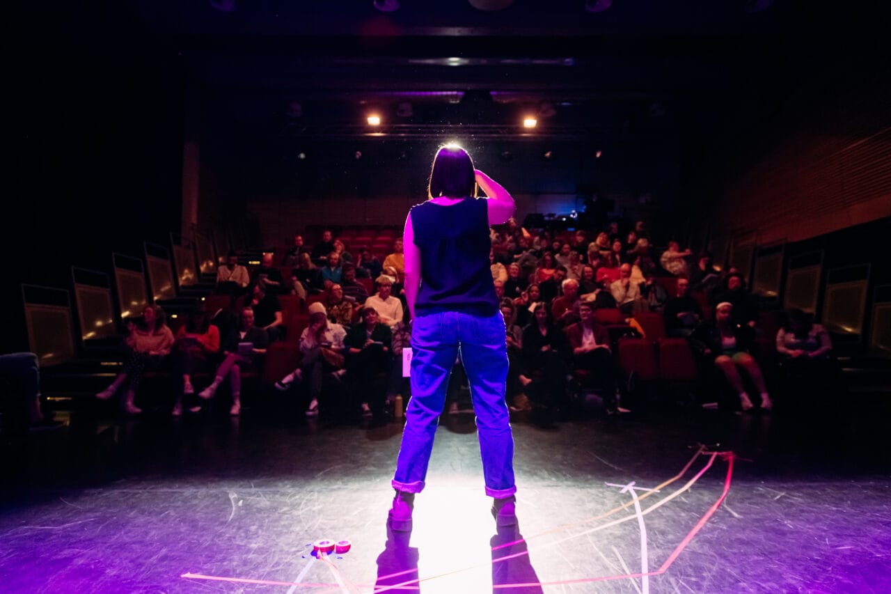 Person standing on a stage facing an audience in a dimly lit theater, with spotlights illuminating the performer from behind.