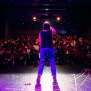 Person standing on a stage facing an audience in a dimly lit theater, with spotlights illuminating the performer from behind.