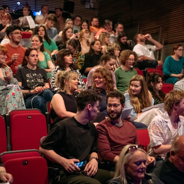 A large group of people sit and talk in a theater with red seats, waiting for an event to begin.