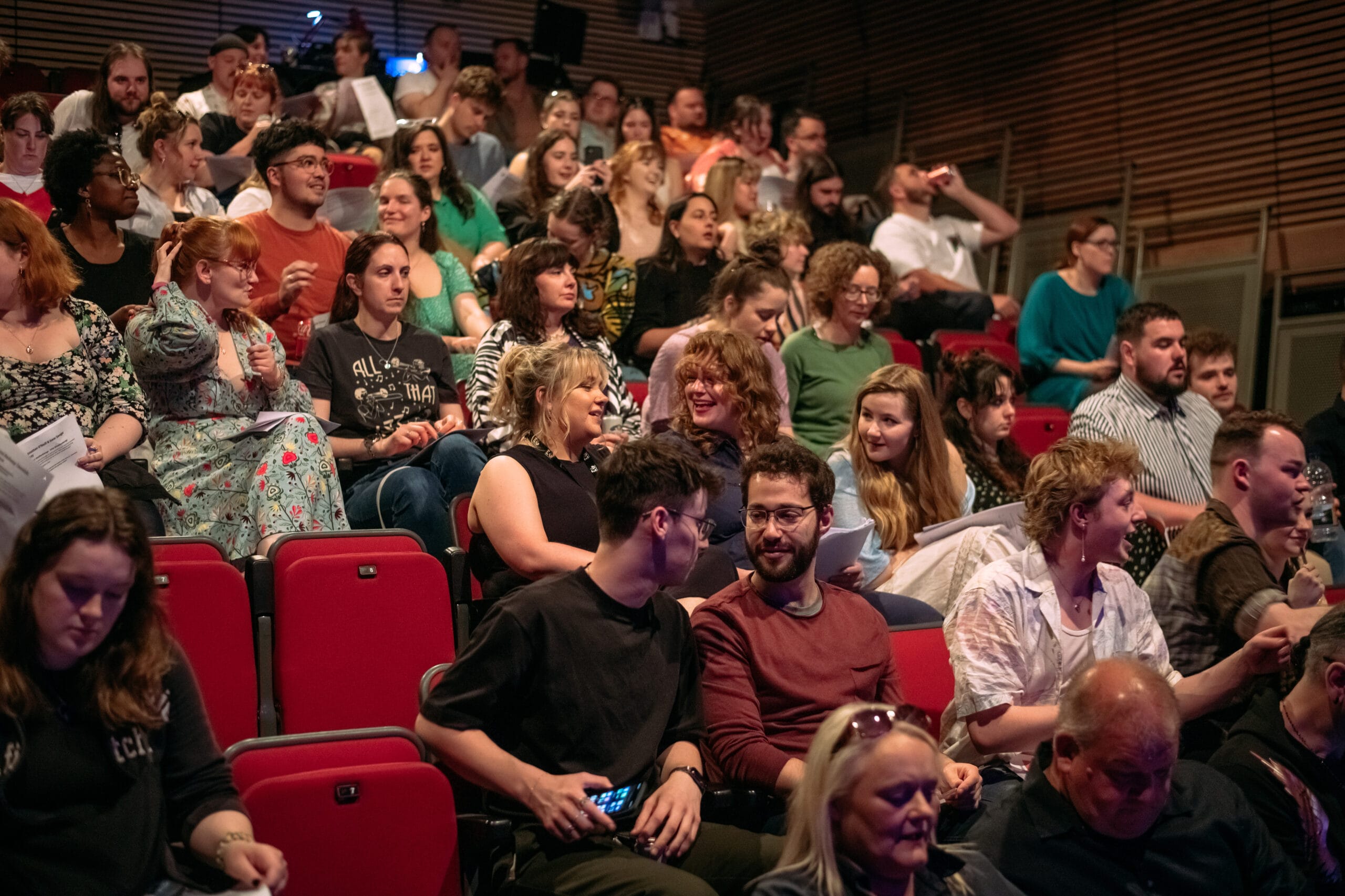 A large group of people sit and talk in a theater with red seats, waiting for an event to begin.