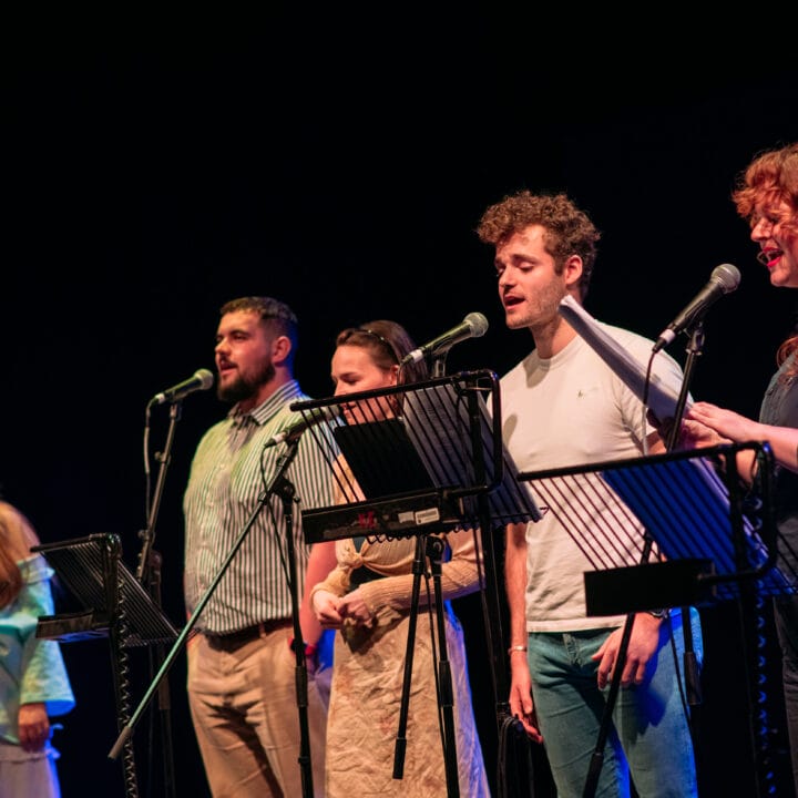 Five people stand in a row, each singing into a microphone and reading from music stands on a dimly lit stage.