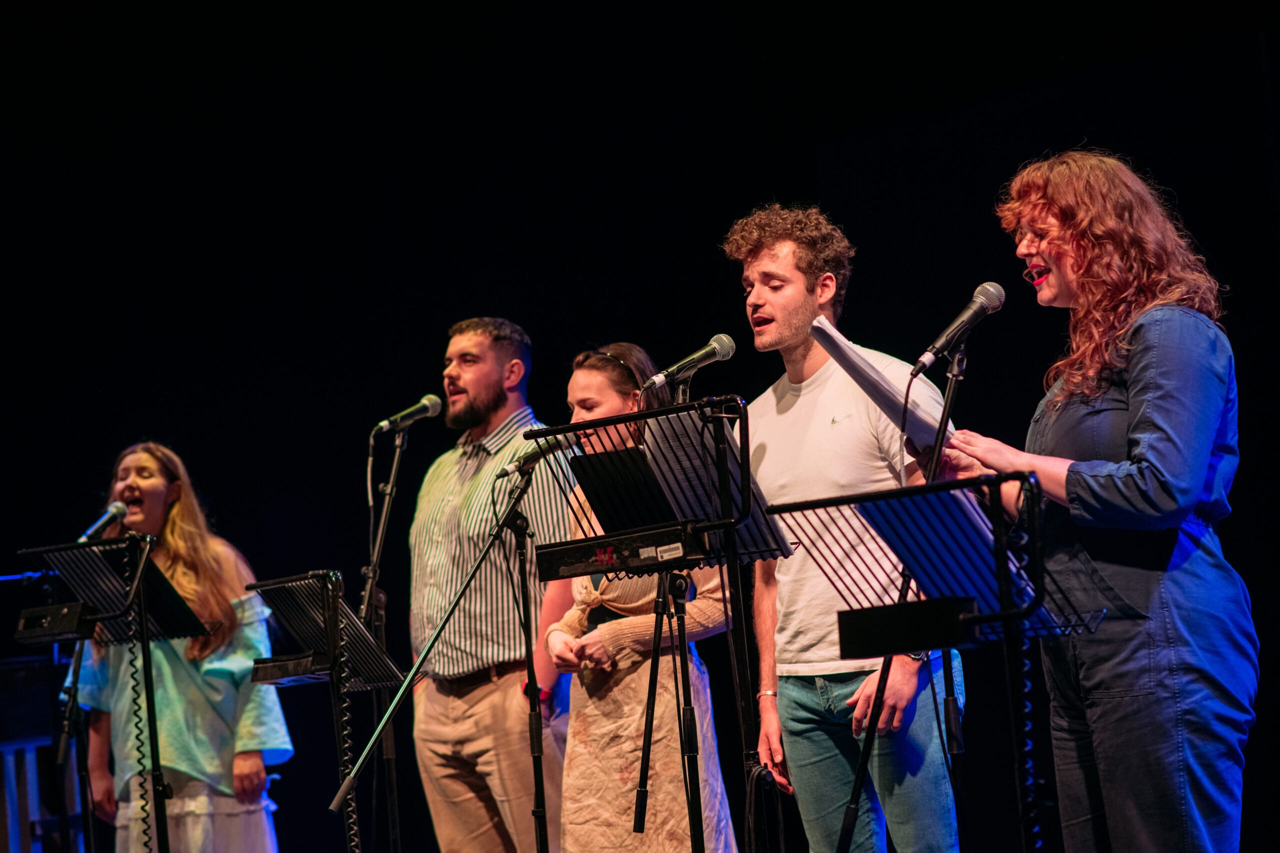 Five people stand in a row, each singing into a microphone and reading from music stands on a dimly lit stage.