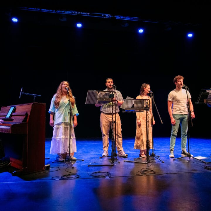 Six performers stand on stage with microphones and music stands, while one person plays a piano on the left; blue stage lights illuminate the scene.