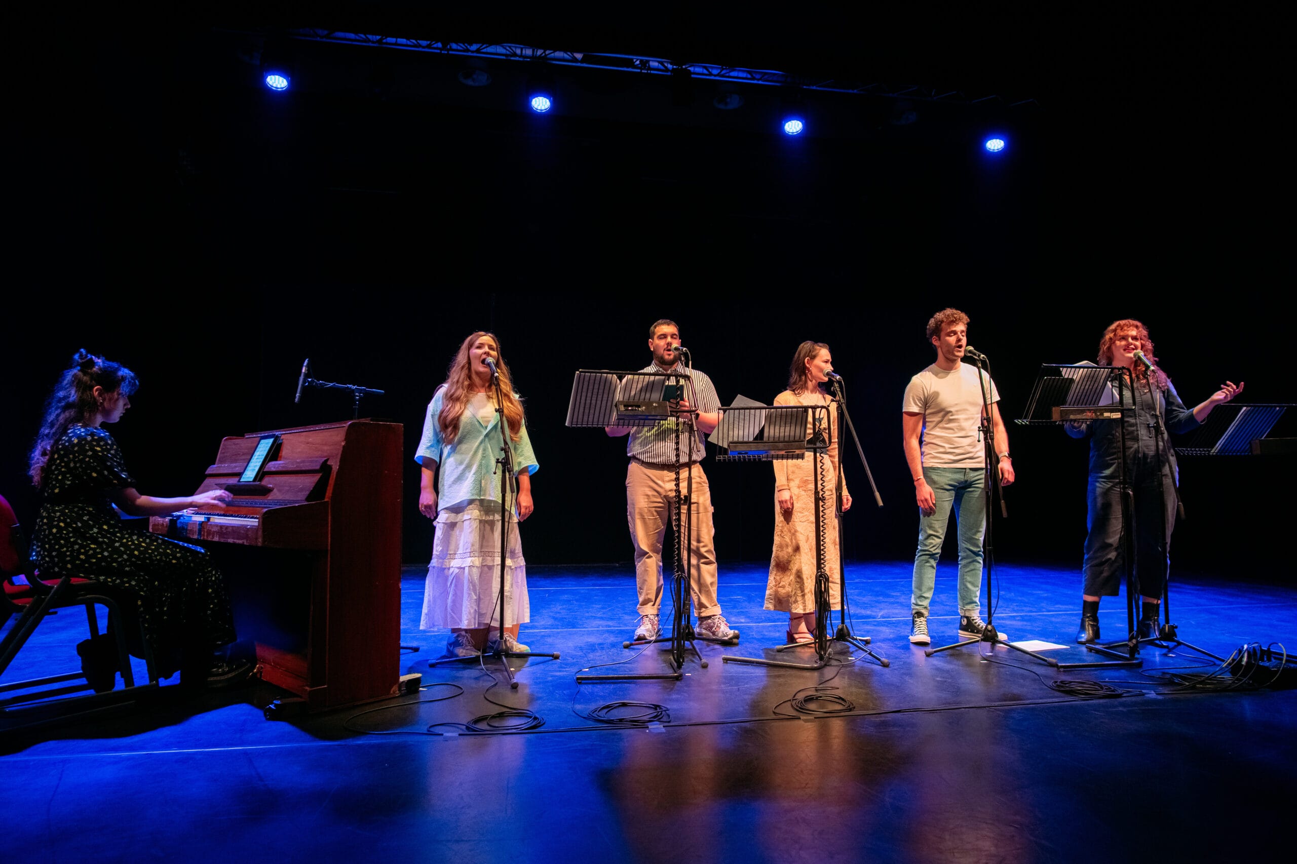 Six performers stand on stage with microphones and music stands, while one person plays a piano on the left; blue stage lights illuminate the scene.
