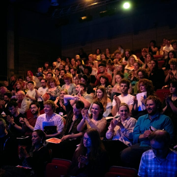 A diverse audience seated in a packed theater applauds a performance, with some people holding programs. The setting is dimly lit with a spotlight overhead.