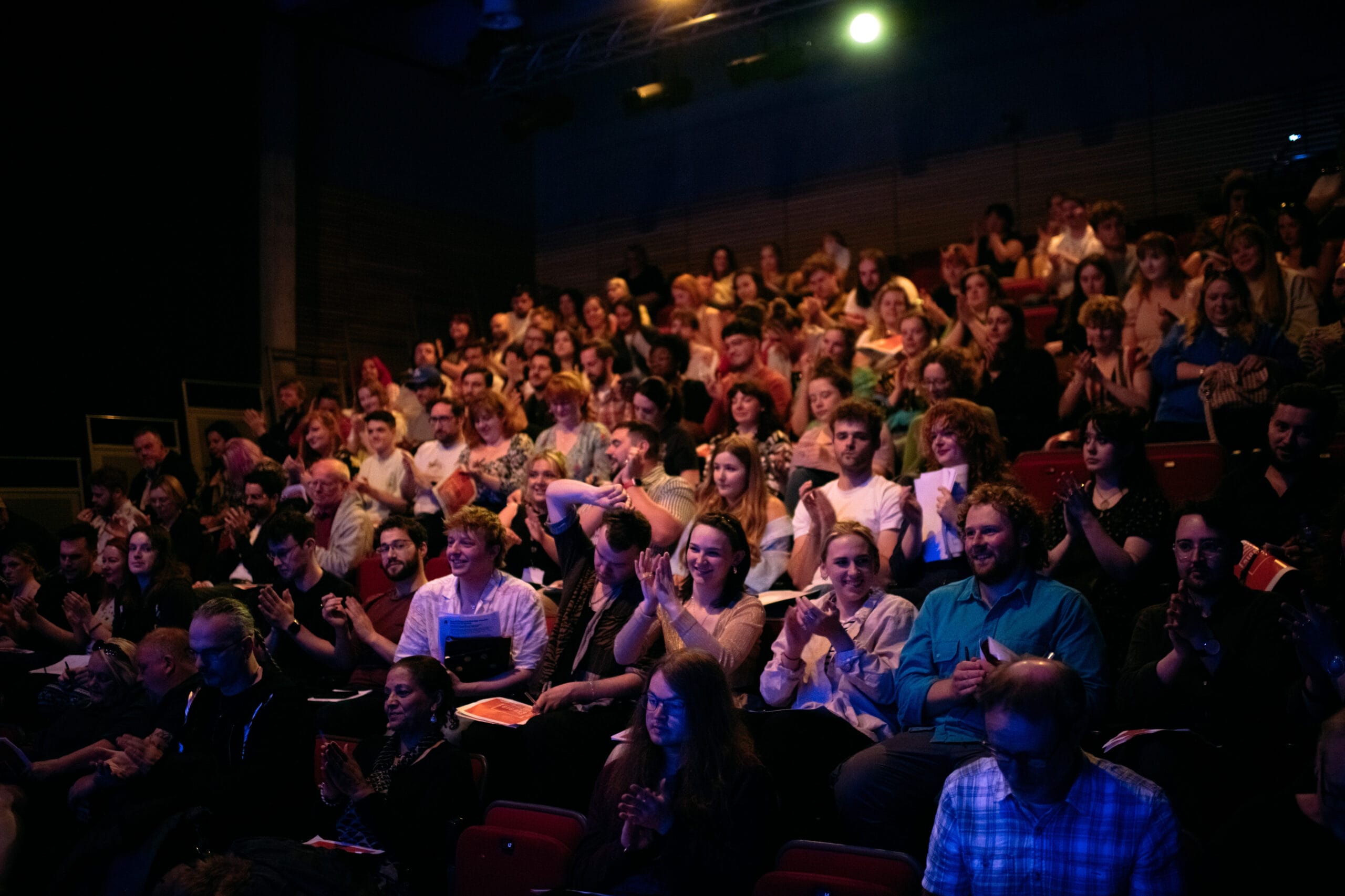 A diverse audience seated in a packed theater applauds a performance, with some people holding programs. The setting is dimly lit with a spotlight overhead.