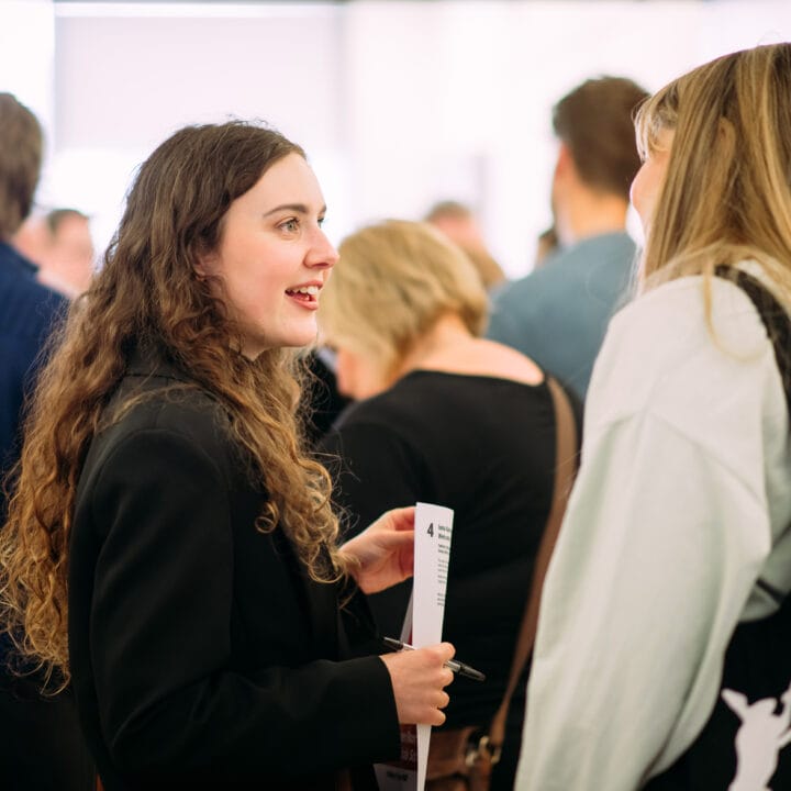 Two women engaged in conversation in a crowded room. One holds a paper and wears a black jacket, while the other has long hair and carries a black bag. People are mingling in the background.