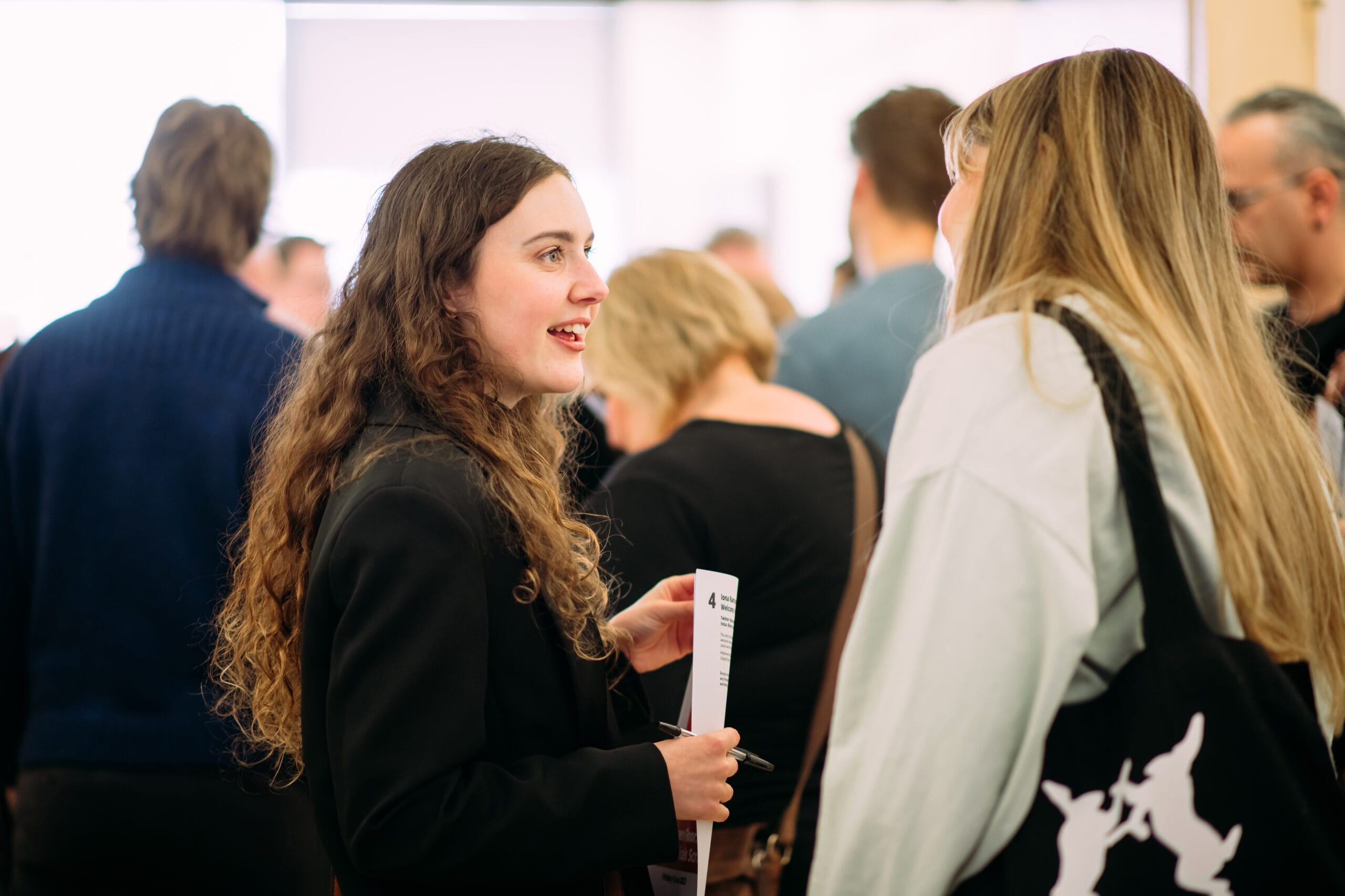 Two women engaged in conversation in a crowded room. One holds a paper and wears a black jacket, while the other has long hair and carries a black bag. People are mingling in the background.