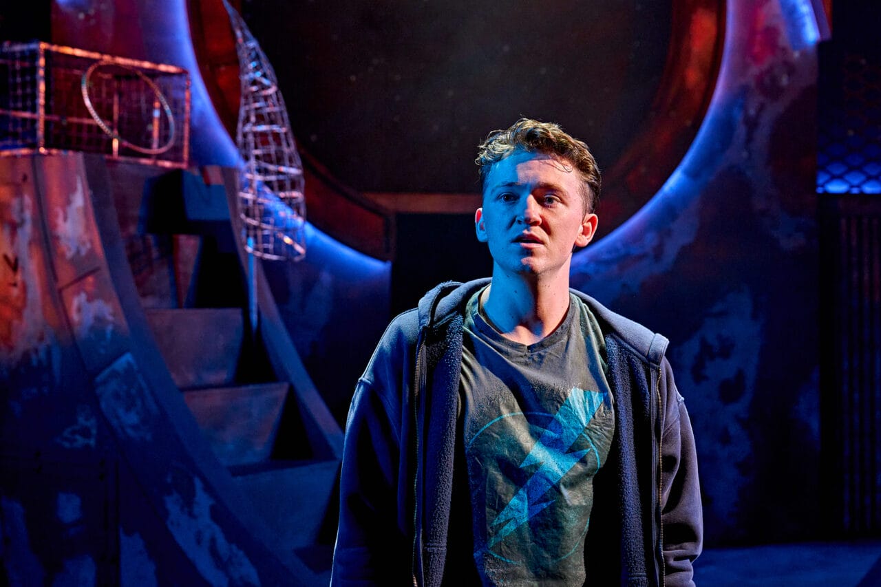 A young man in a t-shirt and hoodie stands under dramatic lighting on a stage with abstract, circular set pieces in the background.