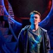 A young man in a t-shirt and hoodie stands under dramatic lighting on a stage with abstract, circular set pieces in the background.