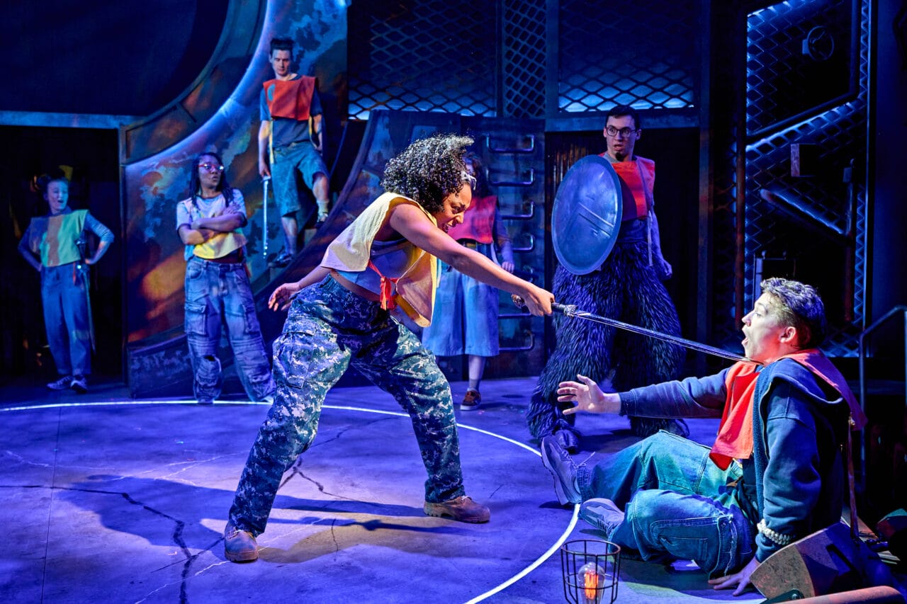 Actors on stage perform a dramatic scene where one person points a sword at another, while others watch in a brightly lit, industrial-themed set.