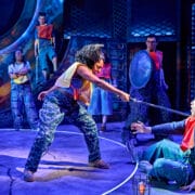 Actors on stage perform a dramatic scene where one person points a sword at another, while others watch in a brightly lit, industrial-themed set.