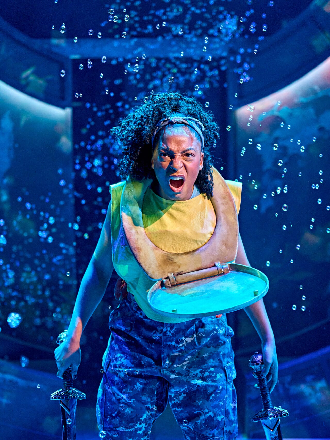 A woman wearing camouflage pants and a toilet seat around her neck shouts while holding two swords on a stage with blue lighting and bubbles in the background.