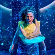 A woman wearing camouflage pants and a toilet seat around her neck shouts while holding two swords on a stage with blue lighting and bubbles in the background.