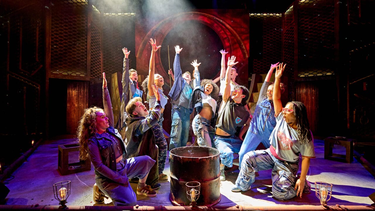 A group of performers on stage, kneeling and standing around a central barrel, reach upward with dramatic lighting highlighting the scene.
