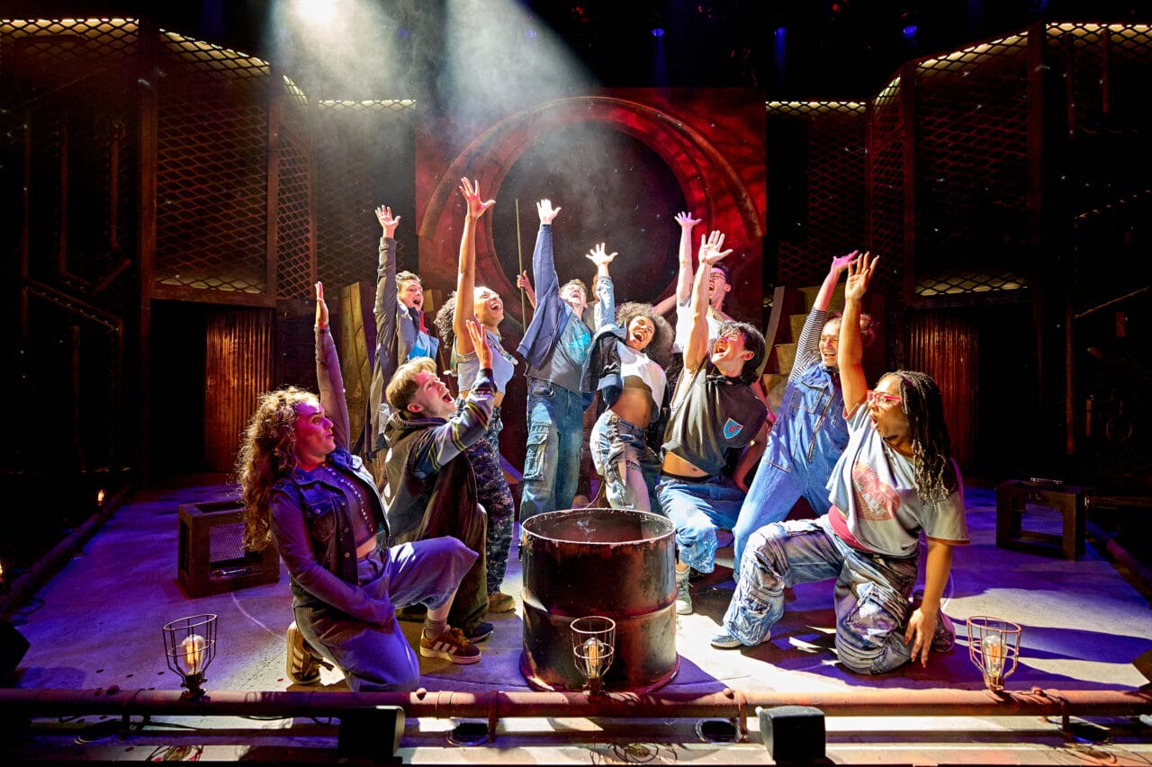 A group of performers on stage, kneeling and standing around a central barrel, reach upward with dramatic lighting highlighting the scene.