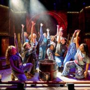 A group of performers on stage, kneeling and standing around a central barrel, reach upward with dramatic lighting highlighting the scene.