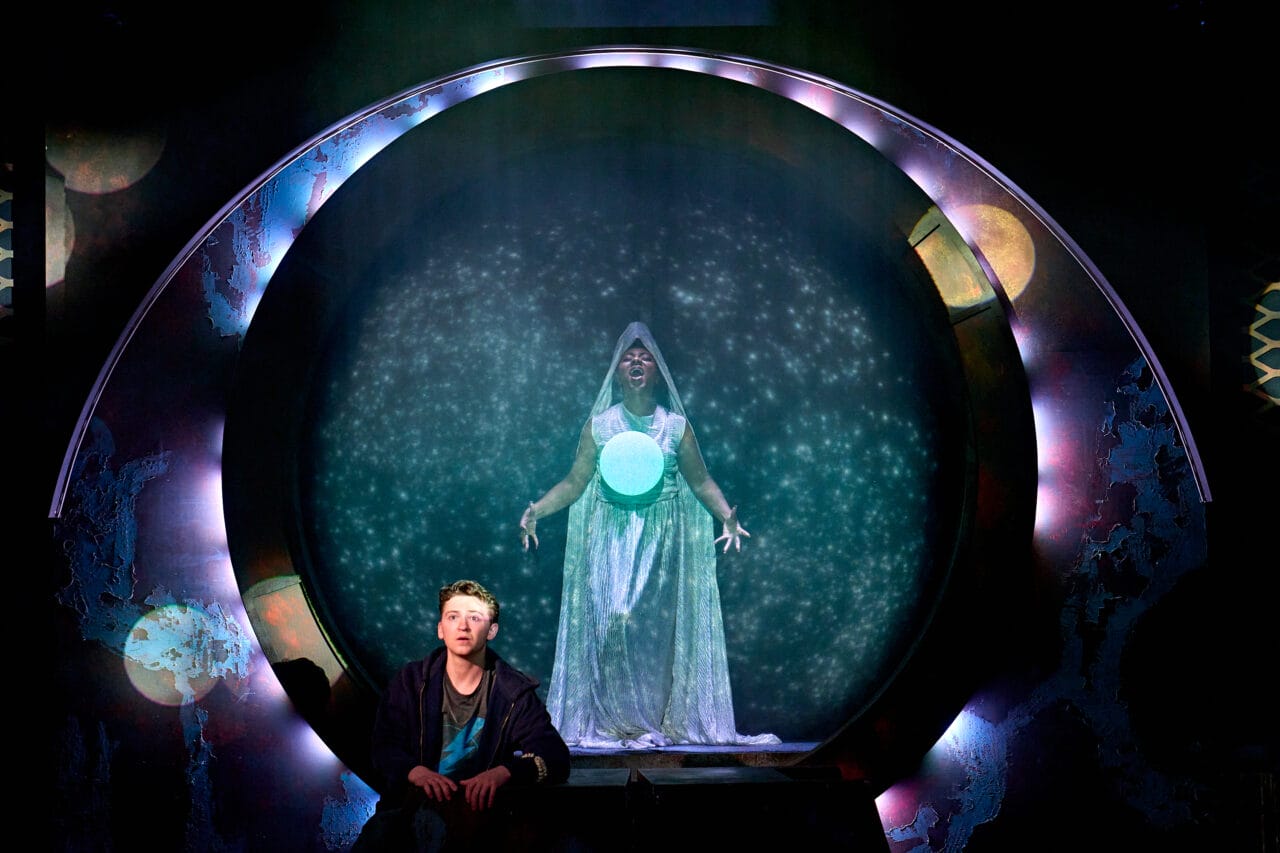 A person in contemporary clothing sits in front of a circular portal, behind which stands a figure in a glowing, hooded robe with a bright orb, surrounded by star-like lights.