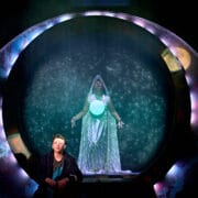 A person in contemporary clothing sits in front of a circular portal, behind which stands a figure in a glowing, hooded robe with a bright orb, surrounded by star-like lights.