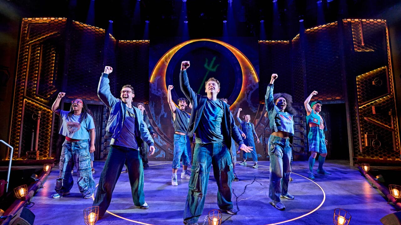 A group of actors on stage perform in front of a large circular set piece, each raising one fist in the air under dramatic blue and orange lighting.