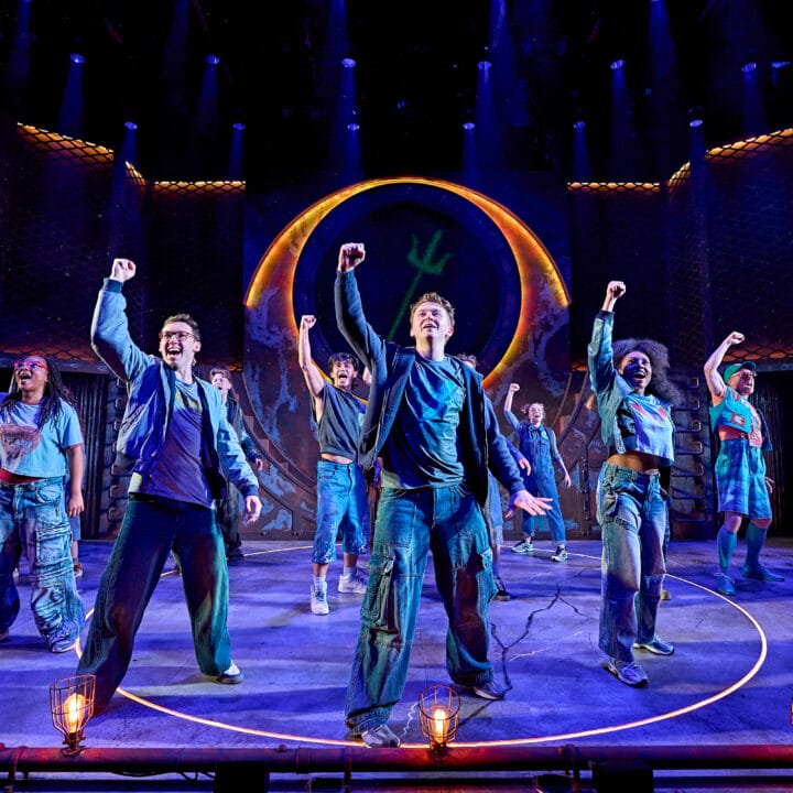 A group of actors on stage perform in front of a large circular set piece, each raising one fist in the air under dramatic blue and orange lighting.