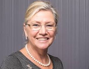 A woman with blonde hair, glasses, and a pearl necklace smiles at the camera against a gray striped background.