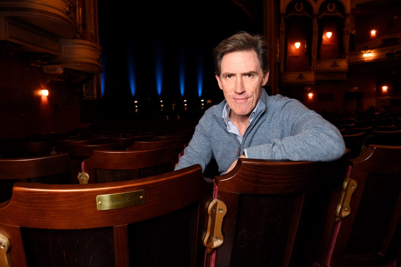 Rob Brydon in a blue zip jumper sits in an empty theatre, leaning on the back of a wooden seat, with the stage and ornate balcony visible in the background.