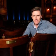 Rob Brydon in a blue zip jumper sits in an empty theatre, leaning on the back of a wooden seat, with the stage and ornate balcony visible in the background.