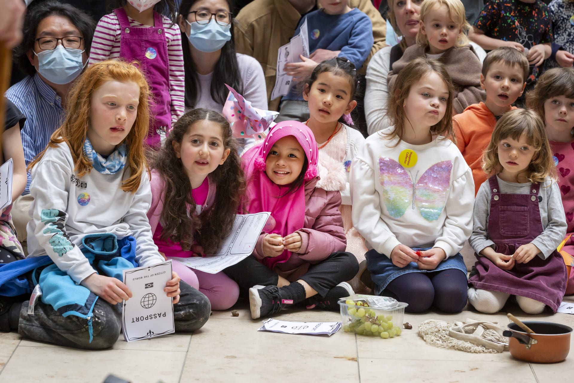 Children sitting on the floor in a crowded room, holding papers and snacks, with adults behind them. Some kids wear colorful clothing and one is wearing a pink hijab.