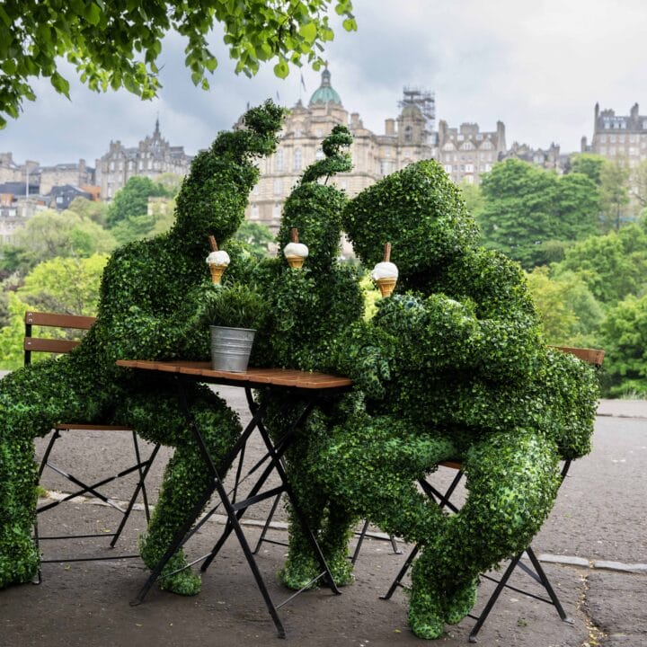 Three people in leafy green plant costumes sit at an outdoor café table, holding ice cream cones, with the greenery of Princes Street gardens in the background.