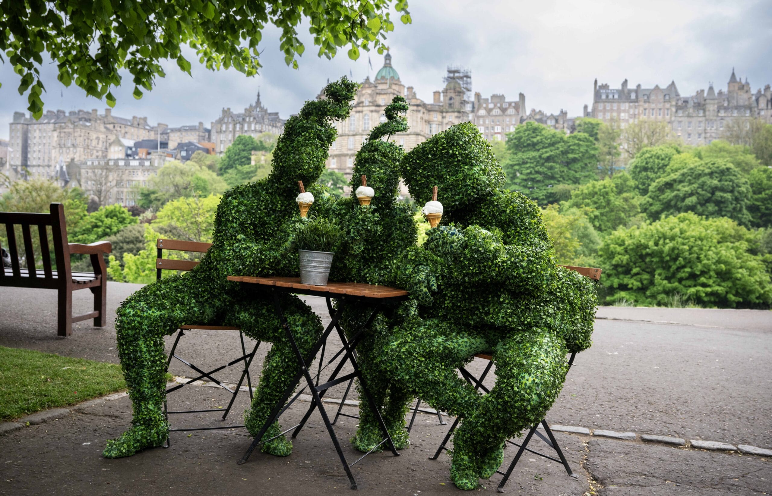 Three people in leafy green plant costumes sit at an outdoor café table, holding ice cream cones, with the greenery of Princes Street gardens in the background.