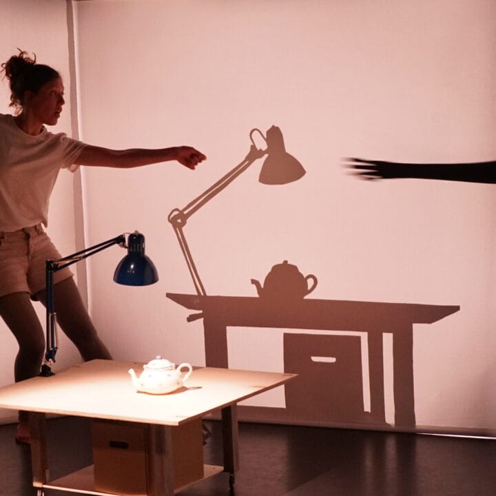 A woman stands next to a desk, reaching toward a lamp. The scene's shadow shows a hand reaching through a tear in the backdrop toward the lamp and teapot.